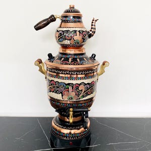 May include: A decorative copper and black samovar with a floral design. The samovar has a matching teapot on top, with a wooden handle and spout. The base is black with colourful lights, and the handles are gold.