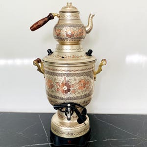 May include: A silver samovar with a matching teapot, both decorated with floral and scrollwork patterns in copper and gold. The teapot has a wooden handle. The samovar sits on a black base. A traditional tea-making appliance.