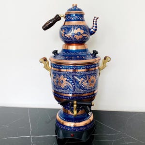 May include: A blue and copper-coloured samovar with floral patterns. The tea kettle sits atop a larger, cylindrical base with handles. The samovar has a wooden handle and a spigot. The base is black.