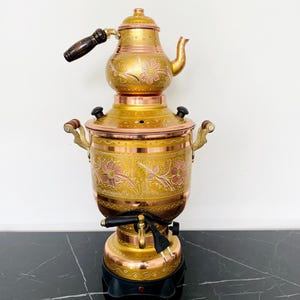 May include: A vintage brass samovar with floral engravings and a copper base. The tea kettle has a wooden handle and a spout. The samovar is on a black base with a power cord. The overall color scheme is gold and copper.