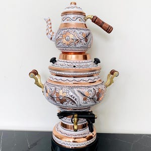 May include: A decorative copper and white tea kettle with floral designs. The kettle has a wooden handle and a black base. The design includes copper accents and intricate detailing, showcasing a traditional aesthetic.
