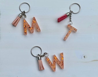 Op de afbeelding: Drie gouden glitterhars lettercharms aan sleutelhangers met roze kwastjes. De letters zijn M, T en W.