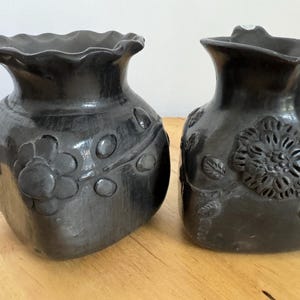 May include: Two black ceramic vases with a glossy finish. The left vase features a scalloped rim and embossed flower and leaf motifs. The right vase has a spout-like rim and a detailed, textured floral relief pattern on its front surface.
