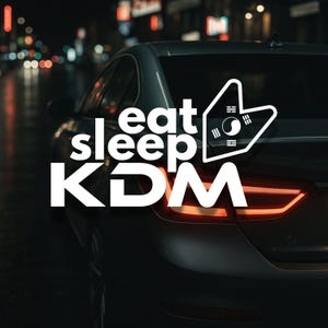 May include: A dark grey car with the words "eat sleep KDM" in white. A white graphic with the South Korean flag is on the car. The car's rear lights are lit, and the background is blurred with city lights.