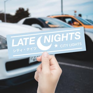 May include: A light blue sticker with white text reading "LATE NIGHTS" and "CITY LIGHTS", plus a crescent moon and star. Japanese text is also present. Cars are visible in the background.