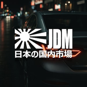 May include: A white JDM (Japanese Domestic Market) decal with Japanese text and a rising sun design is affixed to the rear of a gray car. The car is parked on a wet street at night, with blurred city lights in the background.