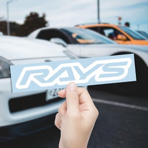 May include: A light blue sticker with the word "RAYS" in white, held in front of a blurred background of cars. The sticker's font is a stylized, bold sans-serif. The sticker is rectangular.