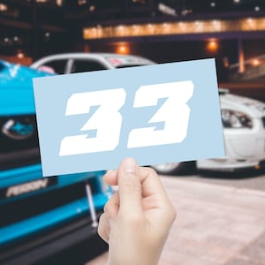May include: A light blue sticker displaying the number "33" in a bold white font. The sticker is held up against a blurred backdrop of cars, hinting at a racing or automotive context.