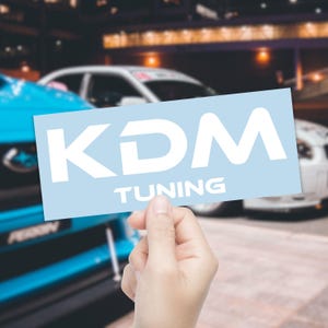 May include: A light blue sticker with the white text "KDM TUNING" is held up in front of a blurred background of cars. The sticker is rectangular and the text is in a bold, sans-serif font.