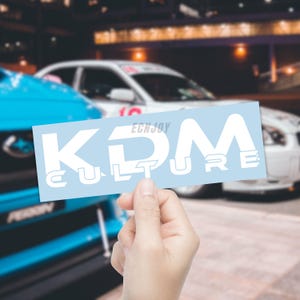 May include: A light blue sticker with the text "KDM CULTURE" in white. The sticker is held in front of a blurred background featuring cars. The word "EGNJOY" is above the "KDM" text.