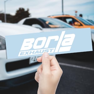 May include: A light blue rectangular sticker with the white text "BORLA EXHAUST". The sticker is held in front of a blurred background of cars. The text is in a bold, sans-serif font.