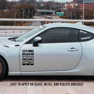 May include: A silver car with black brand logo decals, including Enkei, Mobil 1, Yokohama, Recaro, and HKS. The word "TRUST" is displayed on the rear window. The text "EASY TO APPLY ON GLASS, METAL, AND PLASTIC SURFACES" is also visible.