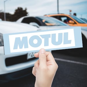 May include: A light blue rectangle with the word "MOTUL" in white letters is held in front of a row of cars. The cars are white and orange, with a blurred background.