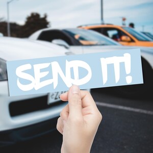 Peut inclure: Un autocollant rectangulaire bleu clair avec l'inscription blanche "SEND IT!". Il est tenu devant une rangée de voitures, avec une voiture blanche au premier plan. L'arrière-plan est flou.