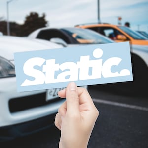 May include: A light blue rectangular sticker with the word "Static" in bold white letters. The sticker is held in front of a blurred background of cars, suggesting an automotive theme.
