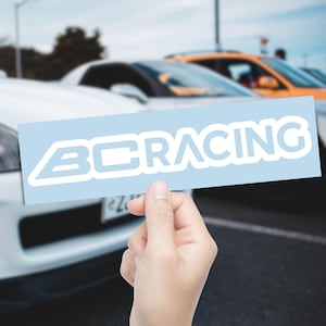 May include: A light blue rectangular sticker with the text "BC RACING" in white. The sticker is held in front of a blurred background of cars, suggesting an automotive theme.