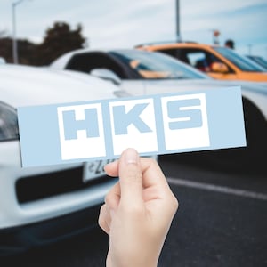 May include: A light blue rectangular sticker with the white letters "HKS" is held in front of a row of cars. The sticker is held by a hand, with the cars in the background slightly out of focus.