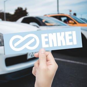 May include: A light blue rectangular sticker featuring the white word "ENKEI" and a white infinity symbol. The sticker is held in front of a row of cars, with a white car in the foreground.