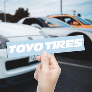 May include: A light blue rectangular sticker with the words "TOYO TIRES" in bold white letters. The sticker is held in front of a blurred background of cars, including a white sports car and an orange vehicle.