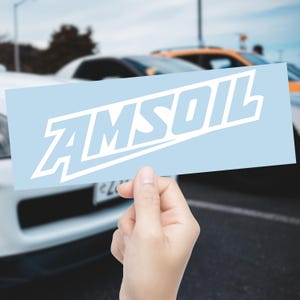 May include: A light blue rectangular sticker with the word "AMSOIL" in white, held in front of a blurred background of cars. The text is in a stylized, bold font.