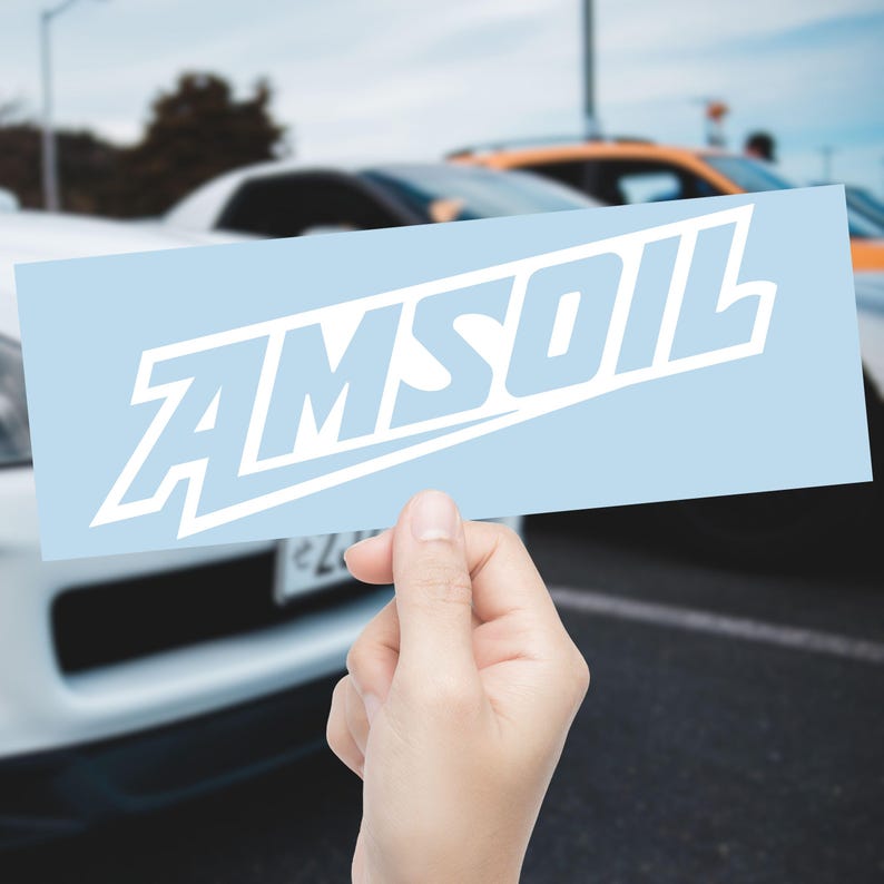 May include: A light blue rectangular sticker featuring the white word "AMSOIL", held up against a blurred backdrop of cars. The text is in a bold, stylized font.