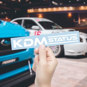 May include: A light blue rectangular sticker with the white text "KDM STATUS" is held in front of two cars. The sticker is in focus, while the cars in the background are slightly blurred.