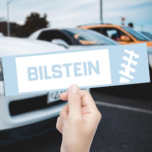 May include: A light blue rectangle with the word "BILSTEIN" in light blue letters. The rectangle is held in front of a row of cars. The background is blurred, but the cars appear to be parked in a car park.