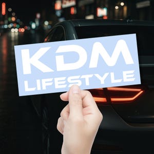 May include: A rectangular light blue sticker featuring the white text KDM LIFESTYLE in a bold, sans-serif font. The sticker is held up against the dark exterior of a car at night, with blurred city lights visible in the background.