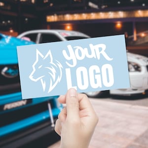 May include: A light blue rectangular sticker featuring a white wolf head logo and the words "YOUR LOGO" in a stylized font. The sticker is held in front of a blurred background of cars and a building.