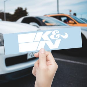 May include: A light blue rectangular sticker featuring the white text "K&N" in a distinctive font. The sticker is held up against a blurred backdrop of cars, hinting at an automotive context.