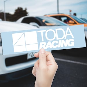 Peut inclure: Autocollant bleu clair avec le texte blanc "TODA RACING". Le sticker présente un motif géométrique. Il est tenu devant une rangée de voitures, avec un arrière-plan flou.