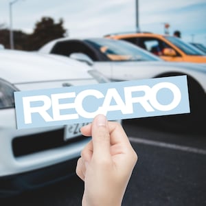 May include: A light blue rectangle with the word "RECARO" in white block letters is held in front of a row of cars. The cars are white and orange, with a blurred background.