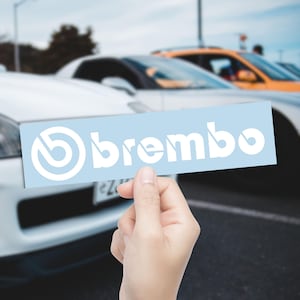 May include: A light blue rectangular sticker with the white text "brembo" and a white logo. The sticker is held in front of a blurred background of cars.