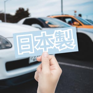 May include: A light blue rectangular sticker with the Japanese characters for "Made in Japan" in white. The sticker is held in front of a row of cars, with a white car in the foreground.