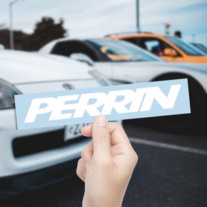May include: A light blue rectangular sticker with the white text "PERRIN" is held in front of a blurred background of cars. The sticker is held by a hand. The cars are white and orange.