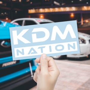 May include: A light blue sticker with the white text "KDM NATION" is held in front of a blurred background of cars. The sticker has a rectangular shape with rounded corners. The text is in a bold, sans-serif font.