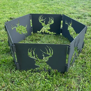 May include: A black metal fire pit with six panels, each featuring a cut-out silhouette of a deer head. The fire pit is assembled in a hexagonal shape and sits on a grassy surface.