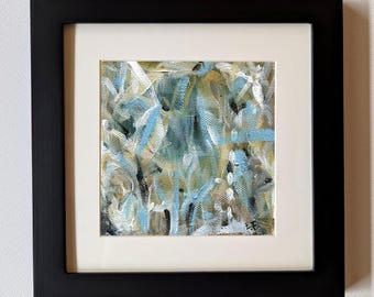 May include: Abstract painting in a black frame. The artwork features a mix of blue, white, and tan brushstrokes. The painting is mounted with a white mat, creating a visual contrast. The framed art is displayed against a white wall.