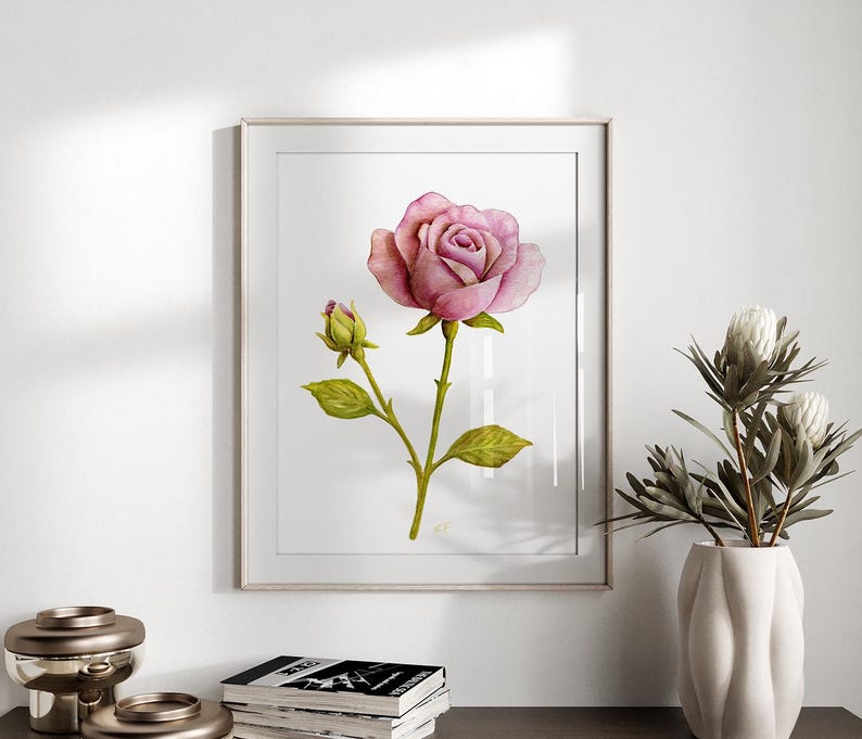 May include: A framed wall art print featuring a watercolor illustration of a single blooming pink rose and a small closed rosebud on a green stem with leaves, centered on a white background within a thin, light-colored rectangular frame.