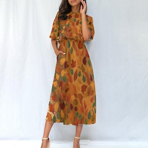 Puede incluir: Vestido midi con estampado de hojas otoñales en tonos naranja, rojo, verde y marrón. El vestido tiene mangas cortas, cintura ceñida y bolsillos. La modelo lleva tacones beige.