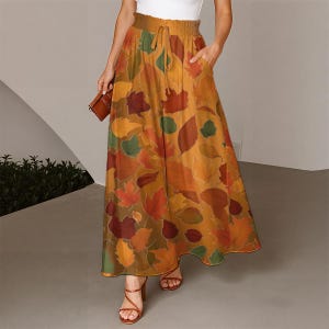 May include: A long, flowing skirt featuring a colourful autumn leaf design in shades of orange, red, and green. The skirt has a drawstring waist and pockets. The model is wearing brown sandals and holding a brown clutch.