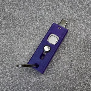 Puede incluir: Una unidad flash USB-C morada con un llavero plateado. La unidad tiene forma rectangular con un botón plateado y un conector USB-C plateado. El fondo es una superficie gris texturizada y brillante.