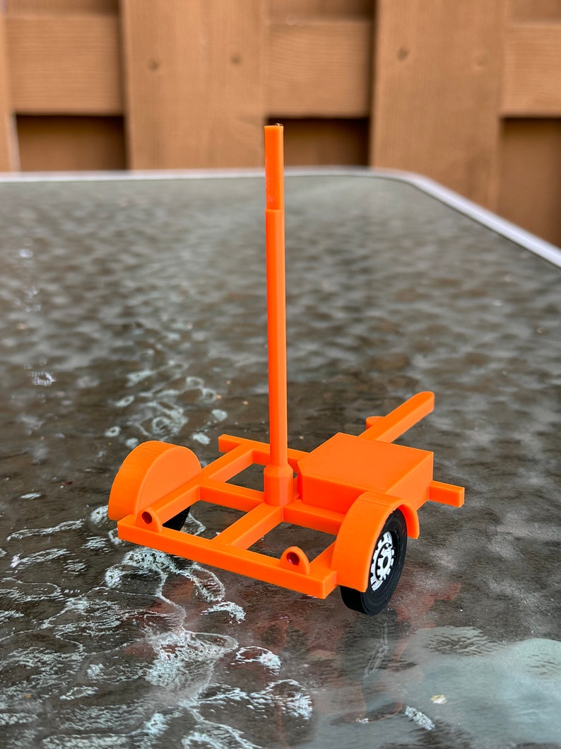 May include: An orange toy trailer with a tall, vertical pole. The trailer has a black wheel on one side and a white wheel on the other side.