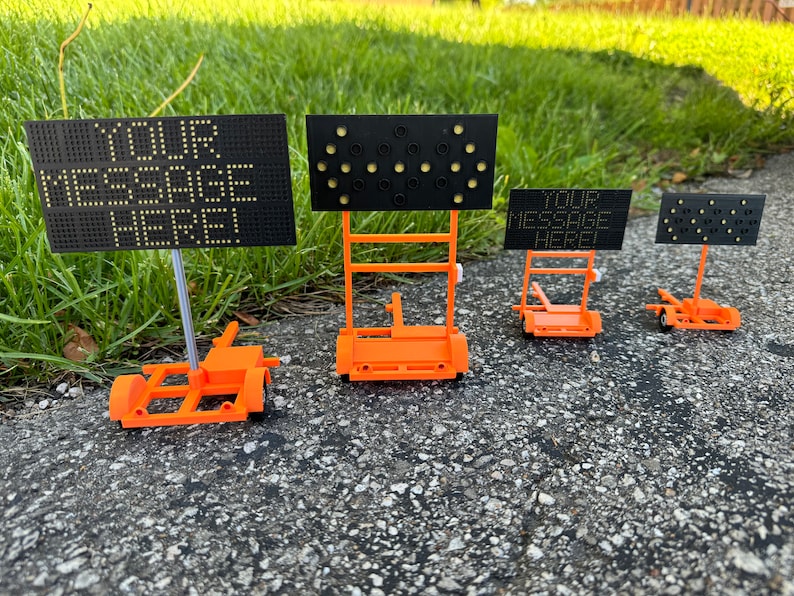 May include: Three orange and black portable message signs with wheels. The signs have different displays, one with the text "YOUR MESSAGE HERE!" and two with blank displays.