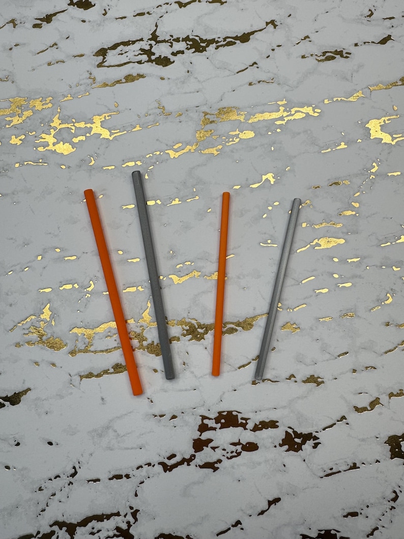 May include: Four drinking straws, two orange and two gray, are arranged on a white surface with gold flecks.