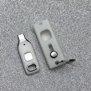May include: A white USB-C flash drive with a silver metallic accent and a small blue indicator light, positioned next to a matching white rectangular protective sleeve featuring a sliding mechanism and a metal key ring attachment.