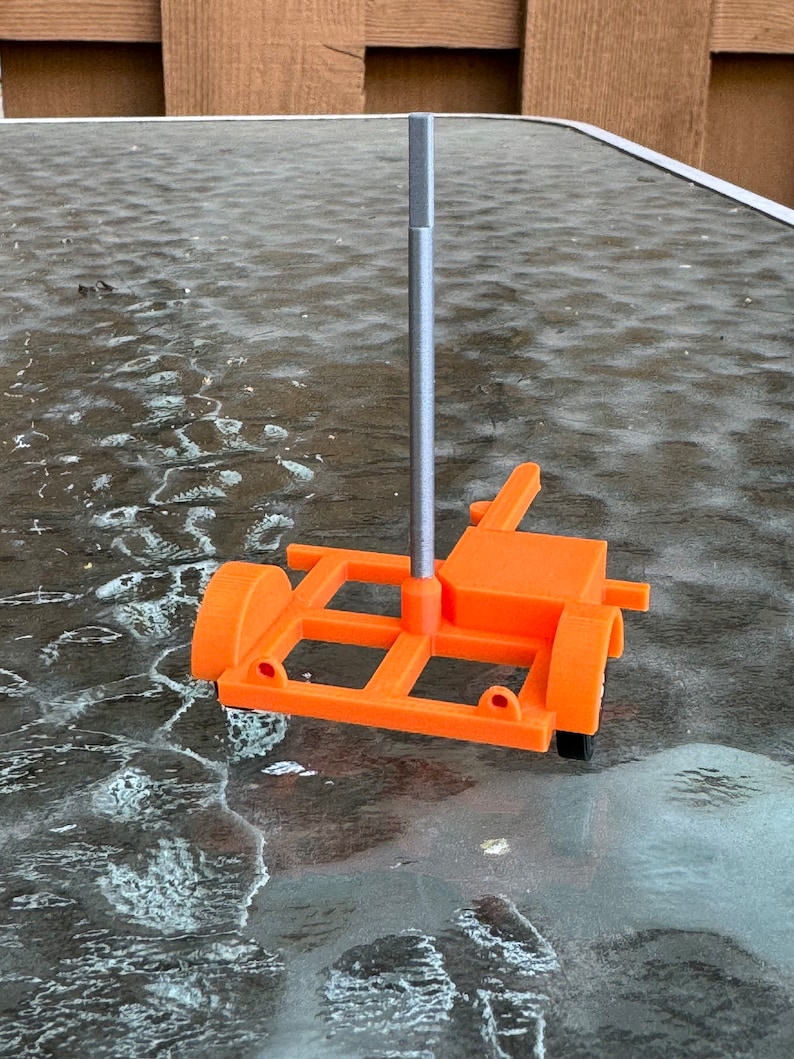 May include: A small orange model trailer with a silver metal pole attached to the top. The trailer has four wheels and a rectangular frame.