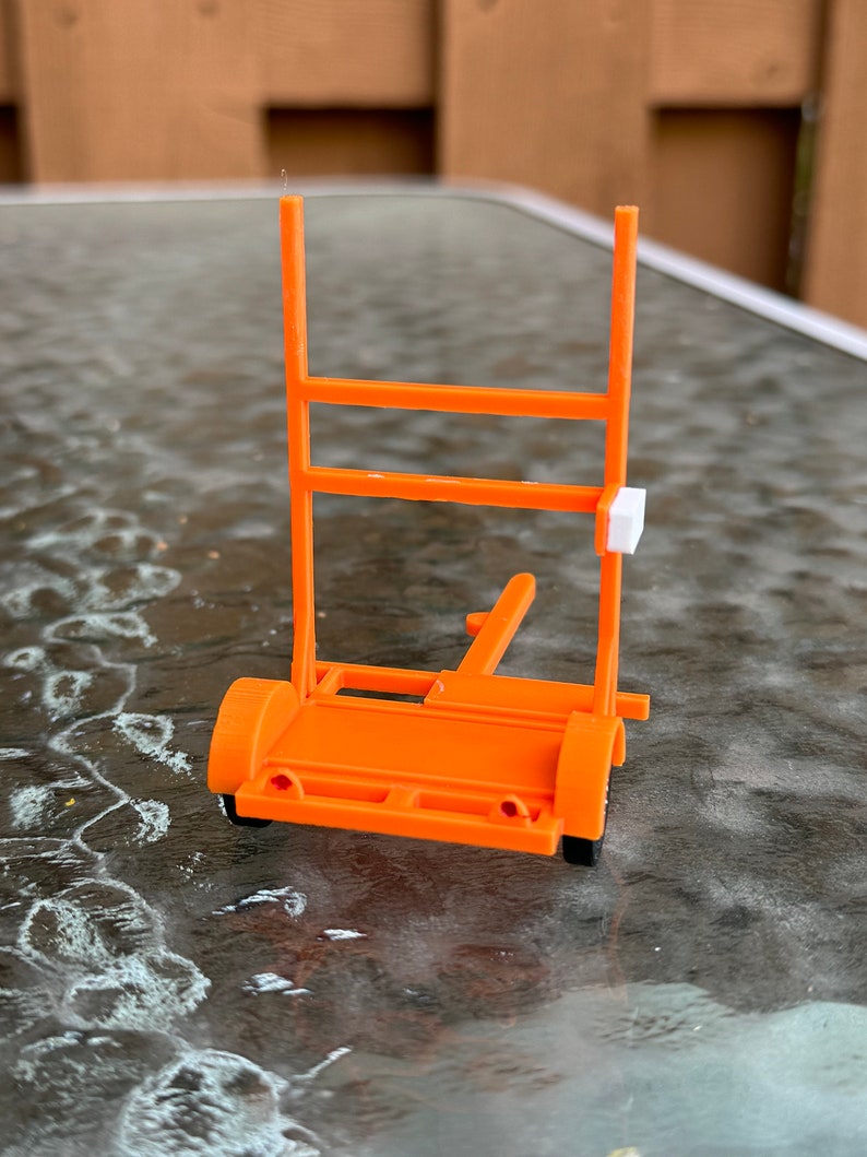 May include: An orange plastic model of a trailer with a white rectangular sign on the back. The trailer has two wheels and a ladder on the back.