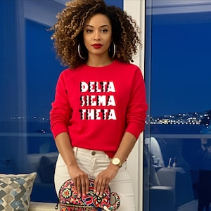 May include: A woman wearing a red sweatshirt with the text "DELTA SIGMA THETA" in white letters. She is wearing white jeans and red ankle boots. She is holding a colorful elephant-shaped clutch.
