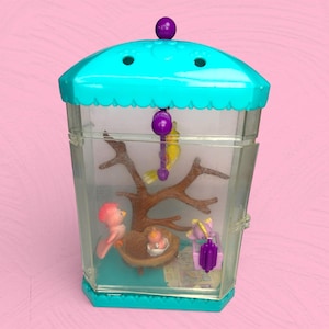 Puede incluir: Un juguete de jaula de pájaros de plástico vintage en tonos turquesa, morado y transparente. La jaula contiene pájaros en miniatura, un nido con un pajarito y una rama de árbol. La jaula tiene un techo festoneado turquesa y una parte superior decorativa morada.